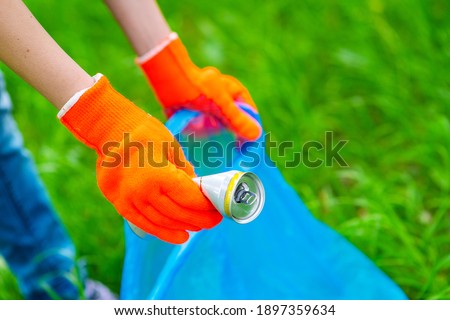 A volunteer’s hand in a bright glove lays a used tin can in a blue plastic bag in a summer park. Rescue of nature concept