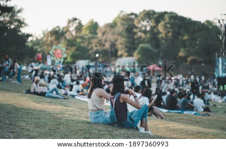 Girls friends watching concert in the park at open air, sitting in front of stage,spectators at background.  Royalty-Free Stock Photo #1897360993