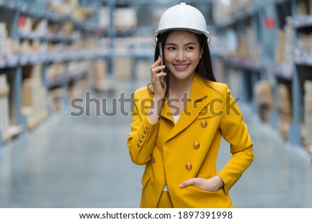 Logistic business storage warehouse shipment. Smart Attractive Asian woman entrepreneur use helmet and mobile phone smile happily to negotiate order with SME customer at warehouse distribution center.