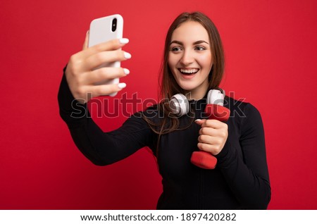 closeup Photo of beautiful smiling positive young brunette woman wearing black sport clothes white headphones nad holding red dumbbell standing isolated over red background using mobile phone taking