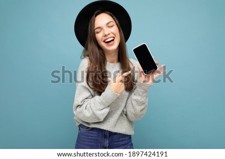 Closeup of charming young happy woman wearing black hat and grey sweater holding phone pointing finger at screen isolated on background