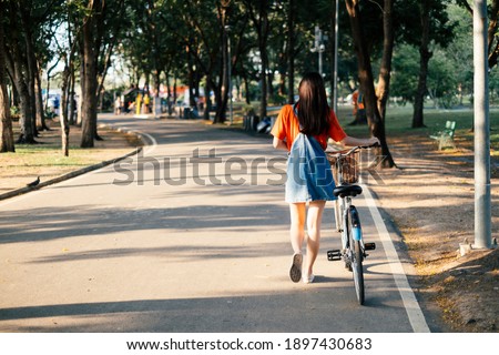 Back shot of long hair girl in orange t-shirt and jeans jumper move her bike along the road in the park.
