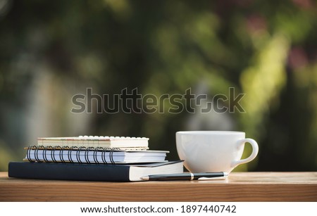 The books and notebook with coffee mug and a pen put on wooden in nature park in the morning atmosphere.