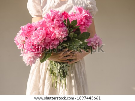 Boho girl holding pink peonies in hands. Stylish hipster woman in bohemian floral dress gathering peony flowers on white backgrounds. International Womens Day. Wedding decor and arrangement