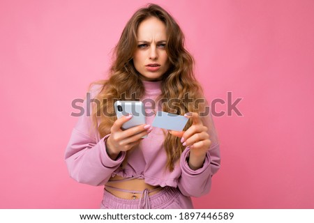 Photo of attractive concentrated young blonde curly woman wearing pink clothes isolated over pink background using mobile phone making payment using credit card looking at camera and having questions