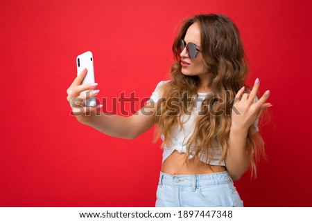 Closeup of happy amazing beautiful young woman holding mobile phone taking selfie photo using smartphone camera wearing everyday stylish outfit isolated over colorful wall background looking at device