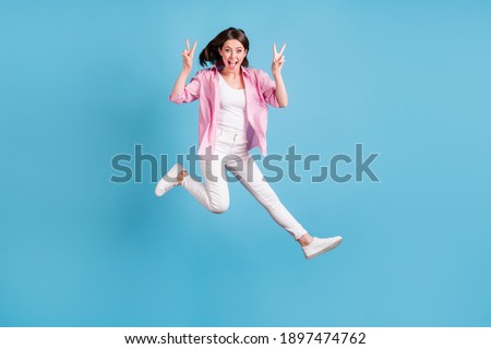 Full size portrait of carefree lady jump show v-sign greetings open mouth isolated on blue color background