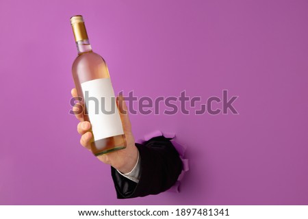 Man's hand holding bottle of wine and breaking through purple paper background.