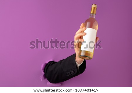 Male hand holding bottle of wine and breaking through purple paper background.