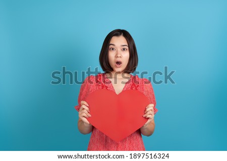 close-up surprised, excited, impressed young asian woman with open mouth in red dress holding big red paper heart isolated on blue background.