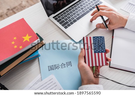 partial view of interpreter holding digital translator with usa flag emblem near paper with chinese hieroglyphs.Translation: 'international contract'