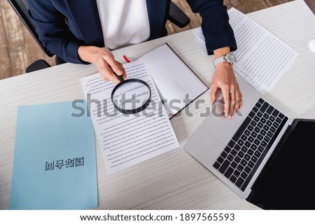 partial view of interpreter holding magnifier near paper with english text and laptop.Translation: 'international contract' Royalty-Free Stock Photo #1897565593