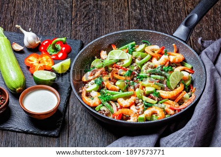 Warm mediterranean salad of lentils, green beans, sweet peppers, shrimps with wilted kale, with lime and tahini sauce dressing served on a frying pan on a dark wooden background, top view, close-up