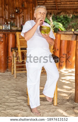 Enjoying the retirement. Happy senior man is drinking a coconut water in the beach bar.