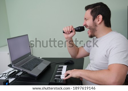 A man blogger, youtuber, influencer recording a video playing the electronic piano and singing at home. Musician, singer, piano player. White screen on laptop.