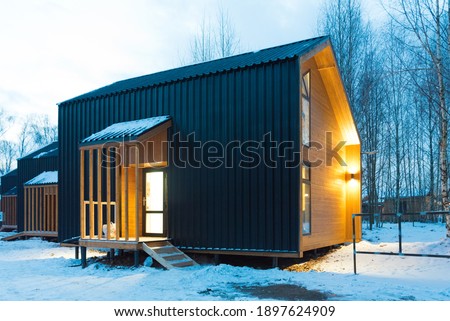 Barnhouse-style homes are surrounded by snow. Evening, warm light shines from the windows. Cozy and calm picture.