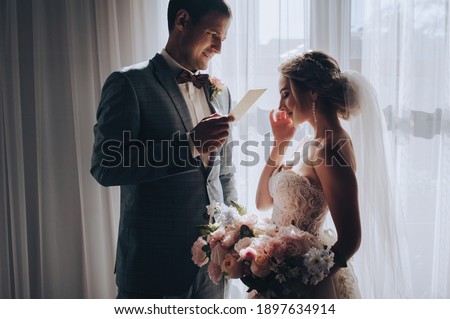 The groom in a checkered suit reads a declaration of love, makes an oath from an envelope, pieces of paper near the window to the bride. Emotional photography. Crying girl wipes tears from her face.