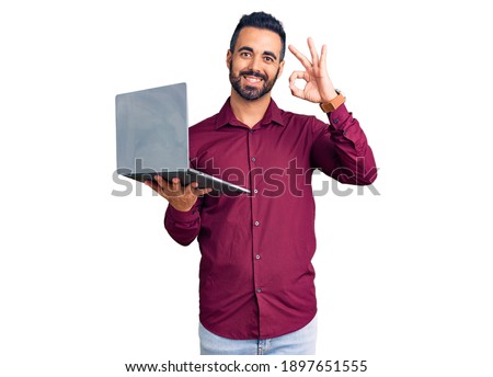 Young hispanic man holding laptop doing ok sign with fingers, smiling friendly gesturing excellent symbol 