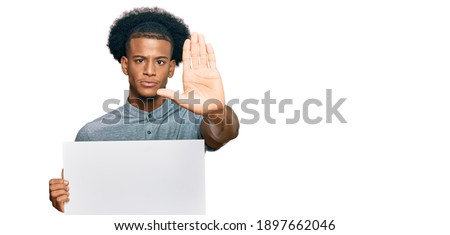 African american man with afro hair holding blank empty banner with open hand doing stop sign with serious and confident expression, defense gesture 