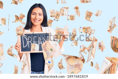 Young beautiful latin girl holding blackboard with dream word smiling happy and positive, thumb up doing excellent and approval sign