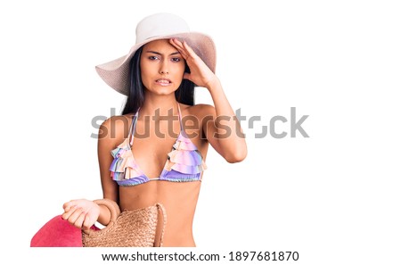 Young beautiful latin girl wearing bikini and hat holding bag stressed and frustrated with hand on head, surprised and angry face 