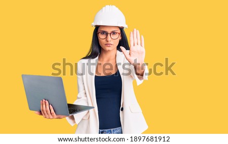 Young beautiful latin girl wearing architect hardhat holding laptop with open hand doing stop sign with serious and confident expression, defense gesture 