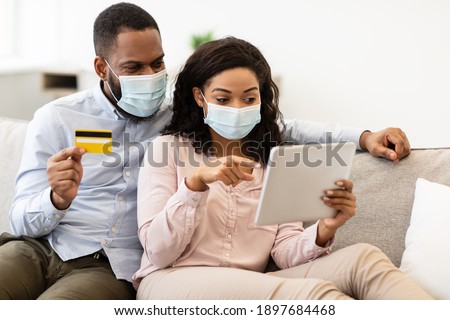 Quarantine Shopping Concept. African american couple wearing disposable protective masks and using digital tablet, man holding credit card. Black family ordering purchases online via internet at home