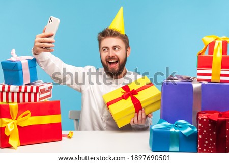 Positive man blogger making selfie on smartphone camera sitting at workplace with many colorful gift boxes around, holidays or birthday celebration. Indoor studio shot isolated on blue background