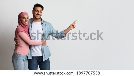 Happy Muslim Couple Embracing And Pointing Aside At Copy Space. Smiling Millennial Arab Spouses Demonstrating Free Place For Your Design Or Advertisement, Islamic Wife Wearing Hijab, Panorama Royalty-Free Stock Photo #1897707538