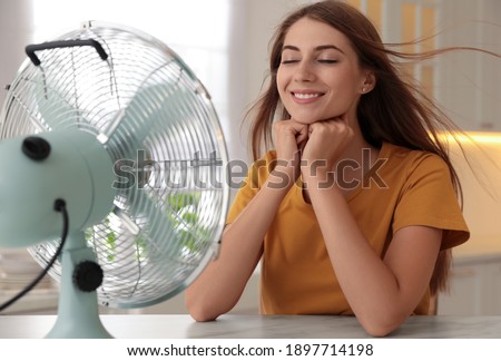 Woman enjoying air flow from fan at table in kitchen. Summer heat Royalty-Free Stock Photo #1897714198