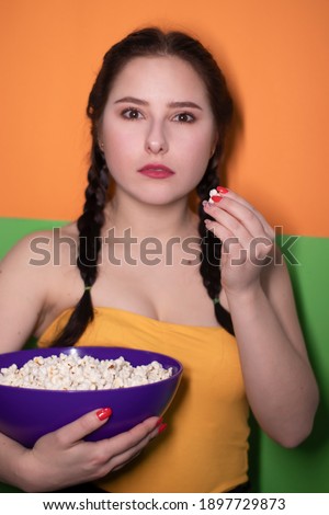 charming brunette woman in yellow top holding a purple bowl with popcorn on bright green and orange background. movie concept. cinema theme