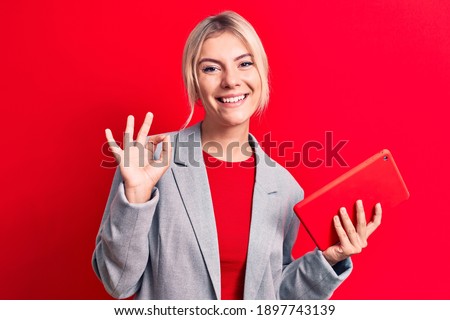 Young beautiful blonde businesswoman working using tablet over isolated red background doing ok sign with fingers, smiling friendly gesturing excellent symbol