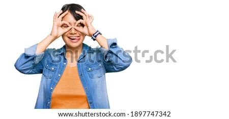 Young beautiful hispanic woman with short hair wearing casual denim jacket doing ok gesture like binoculars sticking tongue out, eyes looking through fingers. crazy expression. 