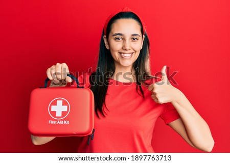 Young hispanic woman holding first aid kit smiling happy and positive, thumb up doing excellent and approval sign 