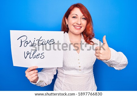 Young redhead woman asking for optimist attitude holding paper with positive vibes message smiling happy and positive, thumb up doing excellent and approval sign