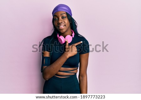 Young african american woman wearing gym clothes and using headphones cheerful with a smile of face pointing with hand and finger up to the side with happy and natural expression on face 