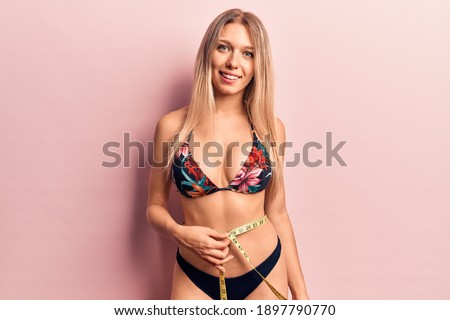 Young beautiful blonde woman wearing bikini using tape measure looking positive and happy standing and smiling with a confident smile showing teeth 