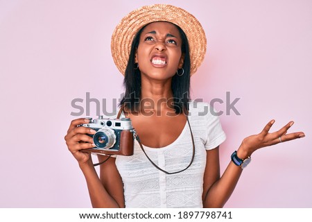 Young african american woman wearing summer hat holding vintage camera crazy and mad shouting and yelling with aggressive expression and arms raised. frustration concept. 