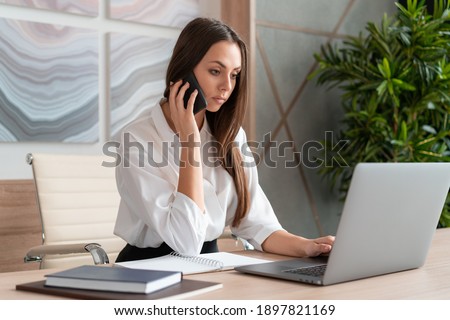 Office manager woman wearing white shirt, sitting to table with laptop making a call looking at the screen, holding a phone. Concentrated office worker in big light office room. Concept of call