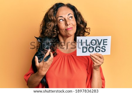 Middle age hispanic woman holding chihuahua dog and paper with i love dogs phrase smiling looking to the side and staring away thinking. 