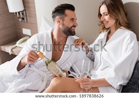 Man in white bathrobe holding bottle of champagne near smiling girlfriend with glasses on hotel bed