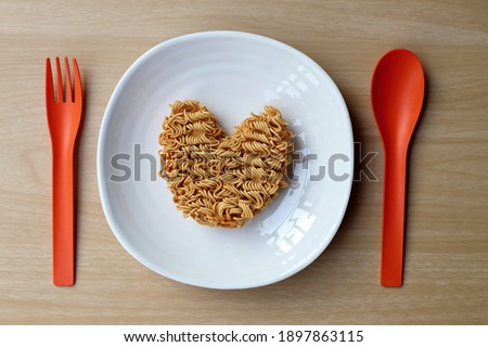 Heart shape of instant noodle in white plate with colorful orange spoon and fork on wooden table, top view.