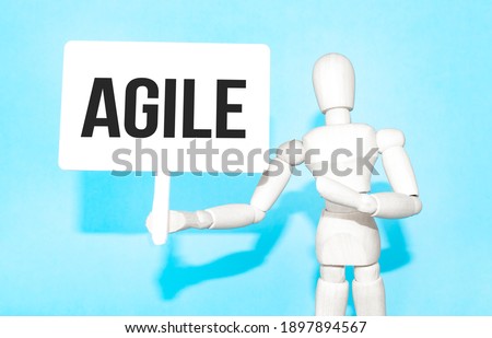 The wooden man holds a white sign with the text AGILE in his hands. The content of the lettering has implications for business concept and marketing.