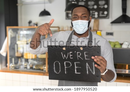 An multiracial waiter, a cafe or bakery owner in protective medical mask is pointing finger at open sign board in his hand. Small business reopening after lockdown