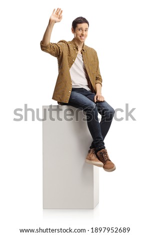 Shy young man sitting on a white column and waving isolated on white background Royalty-Free Stock Photo #1897952689