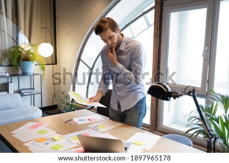 Thoughtful businessman reading documents, analyzing financial project statistics, standing at table in office, pensive executive manager touching chin, pondering developing marketing plan Royalty-Free Stock Photo #1897965178