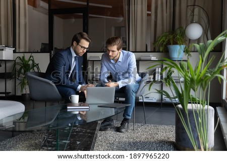 Colleagues using laptop, discussing strategy, sharing ideas, working on project in modern office together, serious mentor coach wearing glasses training new employee intern, explaining task Royalty-Free Stock Photo #1897965220