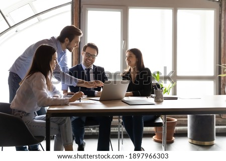 Happy diverse colleagues working on project together, using laptop, sitting at table in modern office, team leader coach training staff, pointing at screen, smiling coworkers discussing strategy Royalty-Free Stock Photo #1897966303