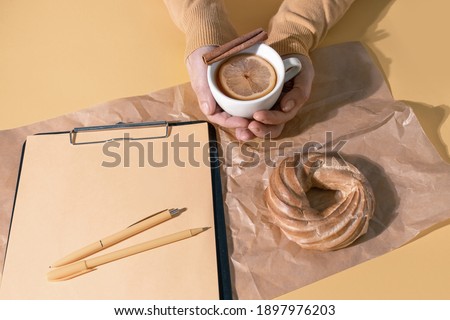 Hands Holding cup of tea or Mulled Wine near Resume Sheet and Cake on the Set Sail Champagne Colour Background, top view.