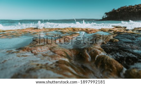 Rocky shore surrounded by calm clear water, small waves and white sand with a clear sky as a background